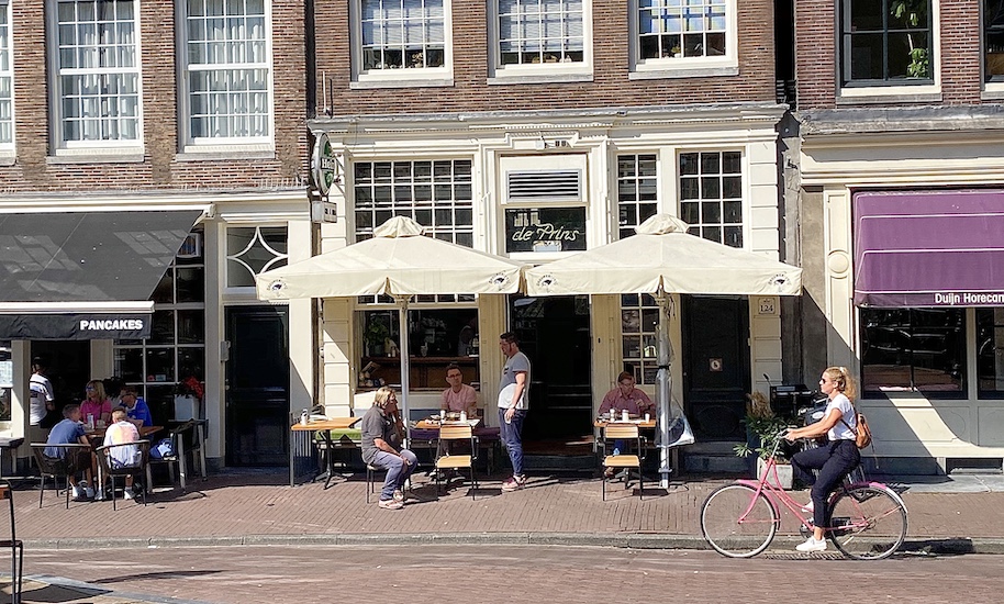 Cyclist rides past people sitting in front of Café de Prins